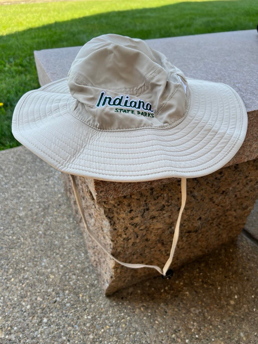 A beige wide-brim sun hat with 'Indiana State Parks' embroidered on the front, resting on a stone block outdoors with grass in the background.