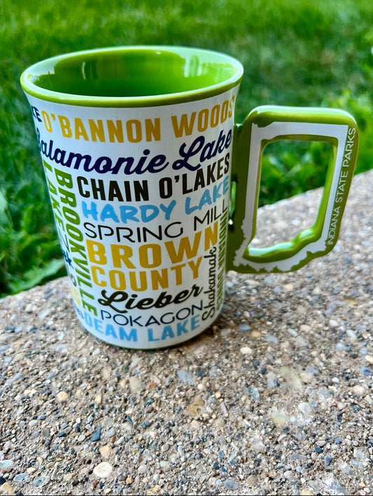 A ceramic mug with a green handle and green interior, printed with various Indiana park names in blue, yellow, and black text on a white background, sitting on a concrete surface with a blurred green lawn behind it.