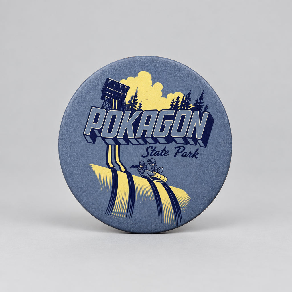 A round magnet showing a toboggan tower, evergreen trees, and two people riding a toboggan down icy tracks, with the text “Pokagon State Park.”