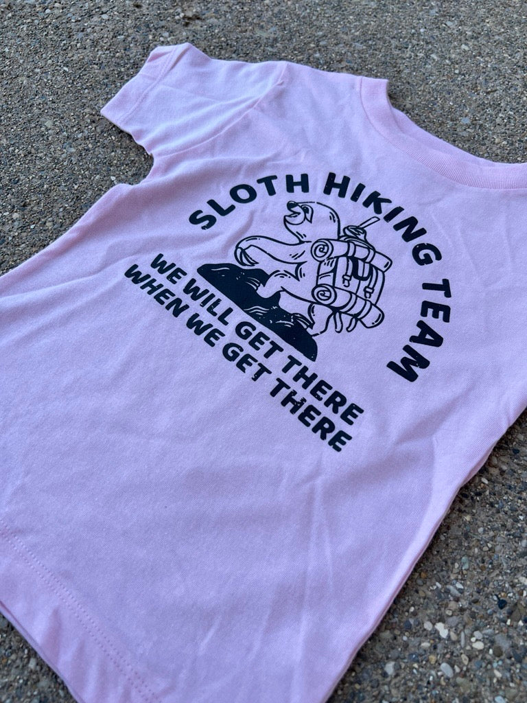 A light pink short-sleeve toddler t-shirt laid flat on a textured gray concrete surface. The shirt features a black graphic of a sloth wearing a backpack, with the text 'SLOTH HIKING TEAM' arched above and 'WE WILL GET THERE WHEN WE GET THERE' printed below.