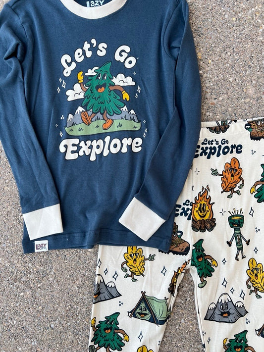 A children's two-piece pajama set laid flat on a textured gray surface. The long-sleeve top is navy blue with white cuffs and collar trim, featuring a cartoon green tree and the words 'Let's Go Explore.