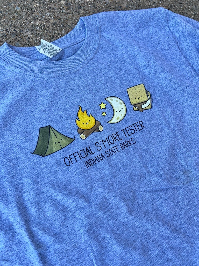 A gray youth t-shirt laid flat on a dark textured surface. The shirt features a cute cartoon graphic of camping and s'mores (tent, campfire, moon, s'more) with the text 'OFFICIAL S'MORE TESTER' and 'INDIANA STATE PARKS' printed below.
