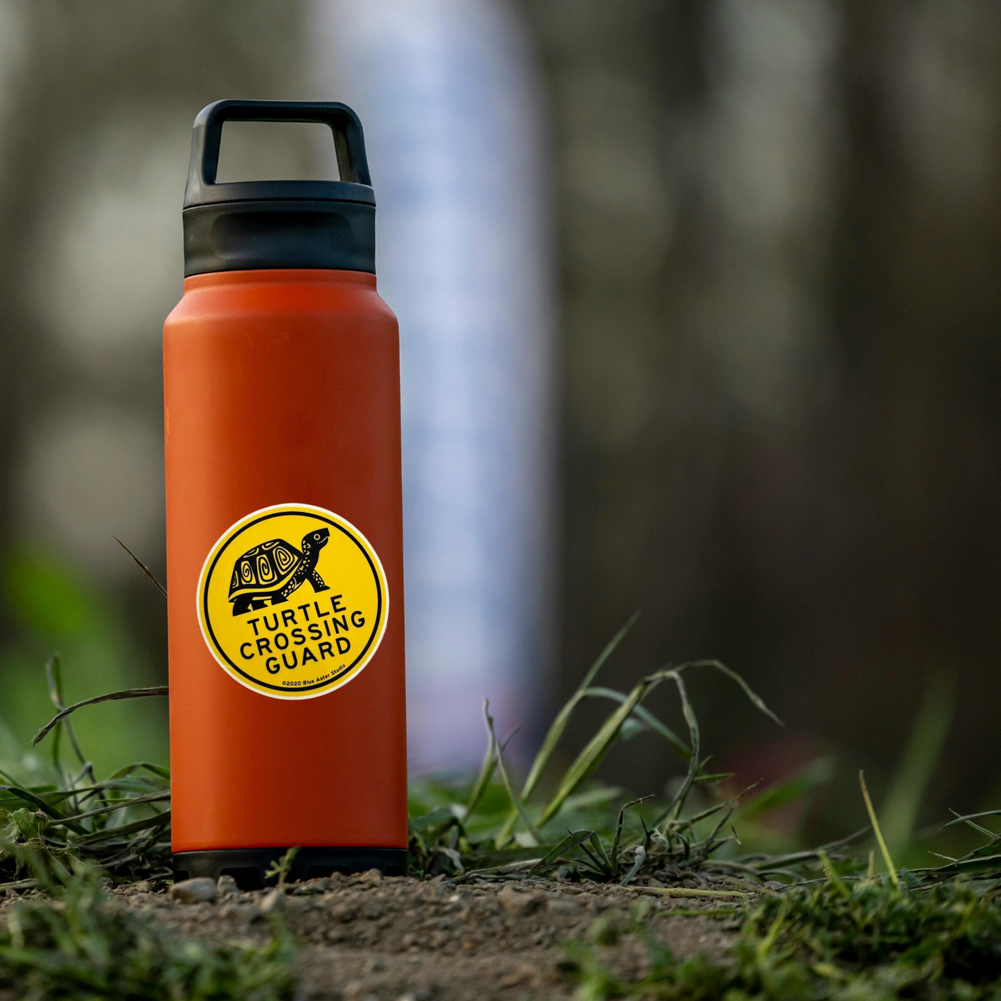 Orange water bottle with a yellow sticker labeled 'Turtle Crossing Guard' on a grassy ground.