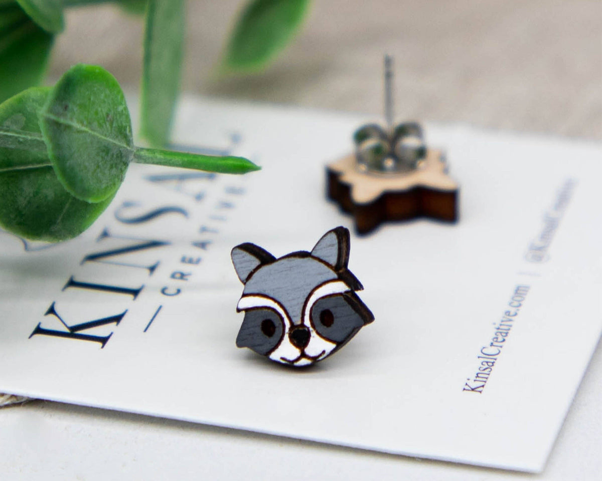 Raccoon-shaped earrings on a Kinsale Creative branded card with green leaves in the background.