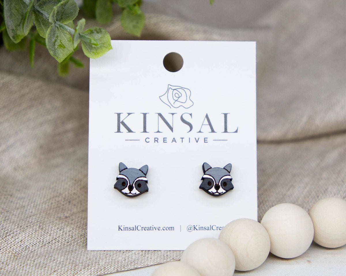 Raccoon-shaped earrings on a Kinsal Creative branded card with decorative elements.