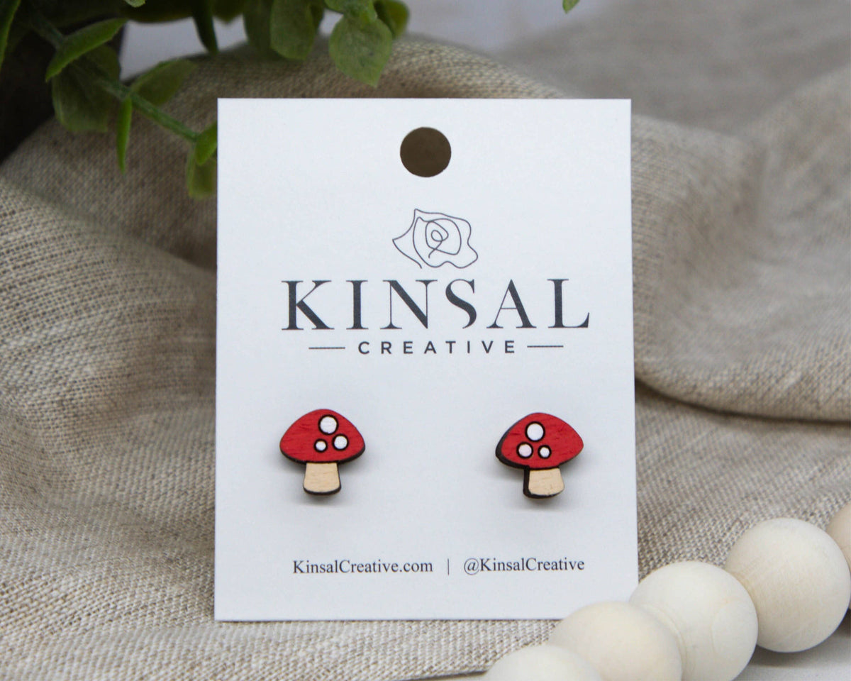Red mushroom-shaped earrings on a Kinsal Creative branded card with a beige fabric background.