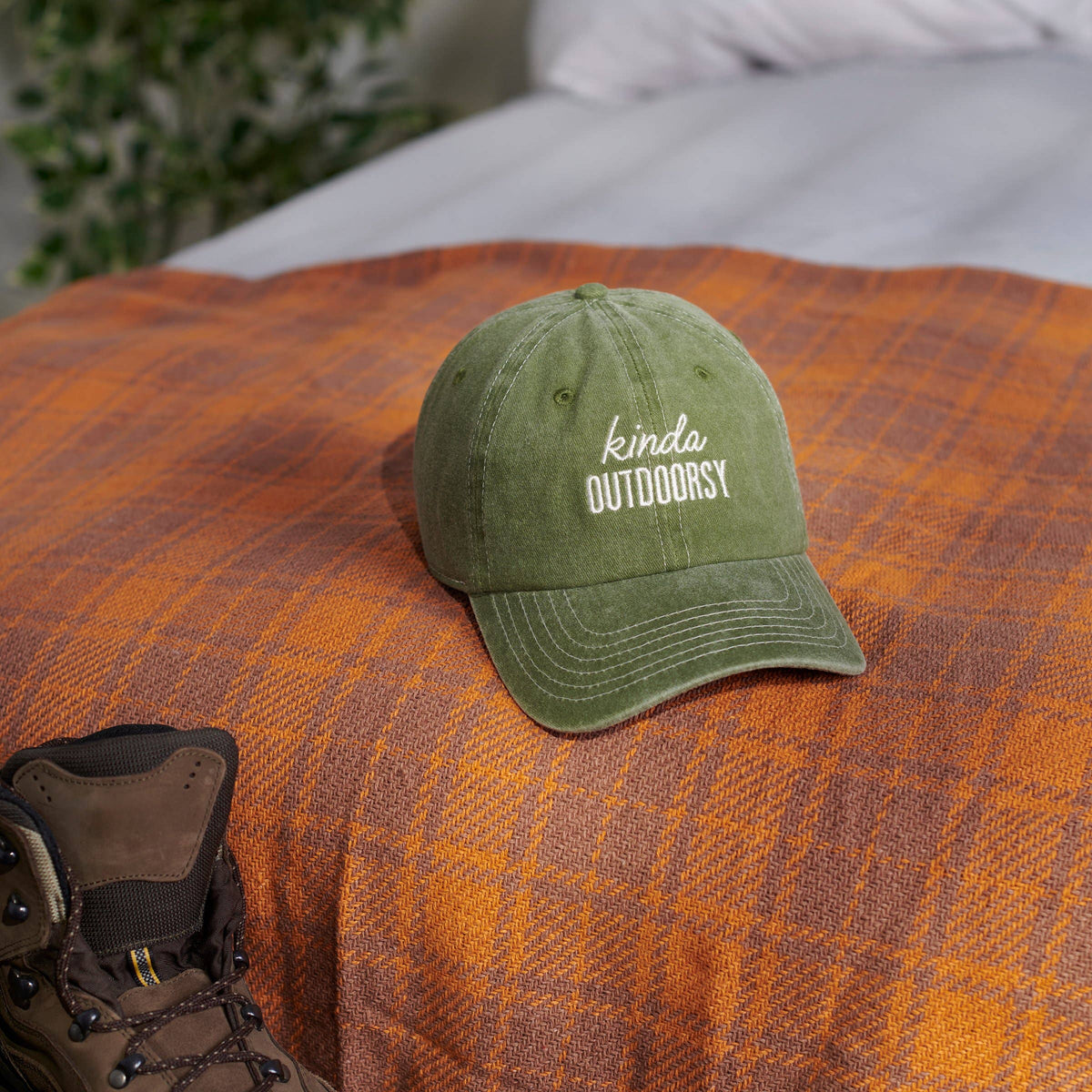 Green cap with 'kinda OUTDOORSY' text on a plaid blanket with a boot nearby.