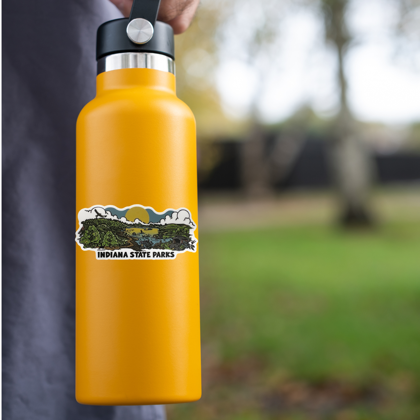 A bright yellow insulated water bottle held by a person outdoors, with a black lid and a printed sticker reading 'Indiana State Parks' on the front. The background is a blurred grassy park area.