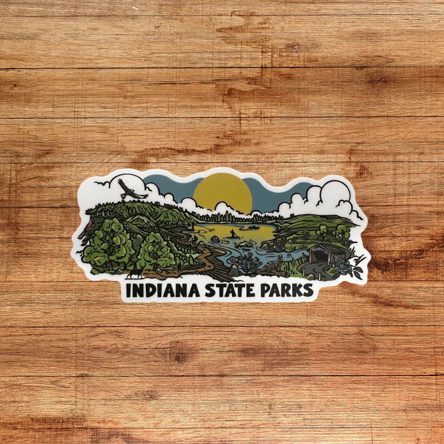 A die-cut sticker featuring an illustrated landscape of Indiana State Parks with green hills, trees, a river, clouds, and a yellow sun, with the text 'INDIANA STATE PARKS' below, shown on a brown wood background.