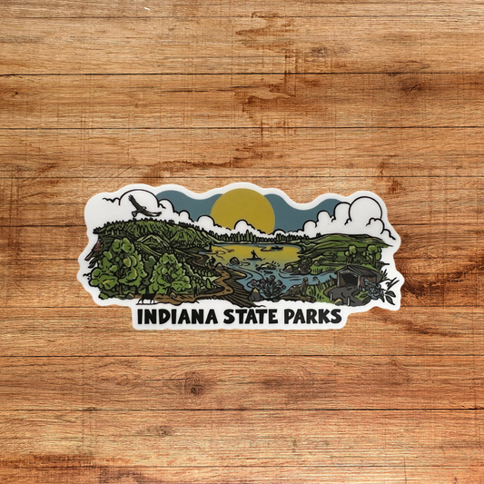 A die-cut sticker featuring an illustrated landscape of Indiana State Parks with green hills, trees, a river, clouds, and a yellow sun, with the text 'INDIANA STATE PARKS' below, shown on a brown wood background.