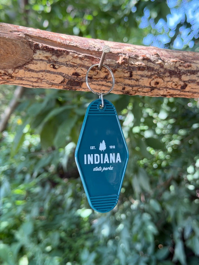 A green Indiana State Parks keychain hanging from a tree branch outdoors, with green foliage blurred in the background. The keychain features Indiana State Parks est 1916 in white text and a silhouette of two trees in white.