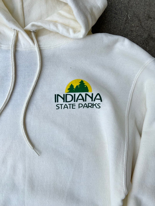 A close-up of a cream-colored hoodie featuring the green and yellow 'Indiana State Parks' logo printed on the left chest, with white drawstrings hanging down.