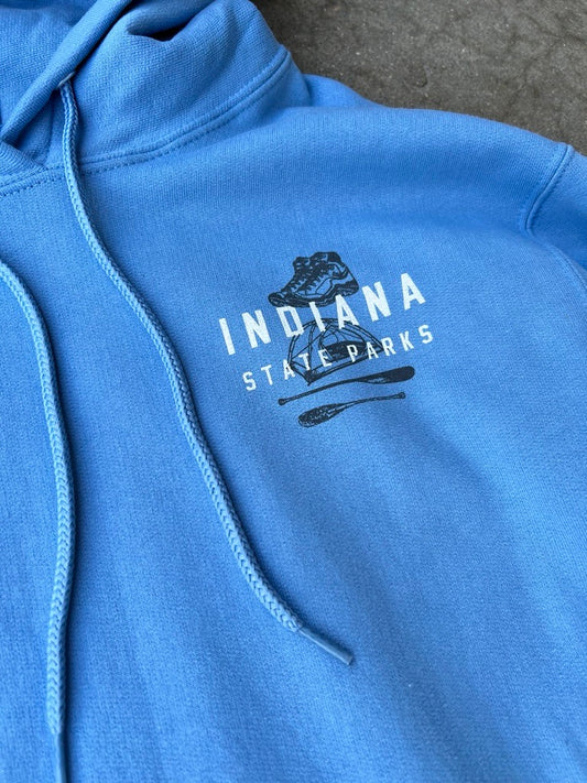 A close-up of a blue pullover hoodie featuring a dark navy graphic on the chest that reads 'INDIANA STATE PARKS' with a small illustration above it, laid flat on a gray textured surface.