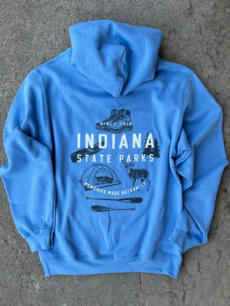 A light blue hooded sweatshirt laid flat on a gray concrete surface, featuring a dark blue graphic print on the back with the text 'INDIANA STATE PARKS' and outdoor-themed illustrations including a hiking boot, tent, deer, and paddleboard.