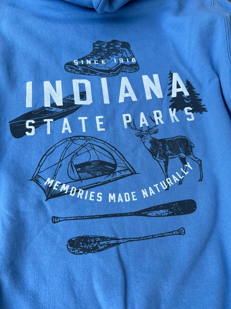 A close-up of a blue hooded sweatshirt featuring a dark navy graphic print for 'Indiana State Parks' with illustrations of a boot, canoe, tent, deer, canoe, and paddles, along with the text 'Since 1916' and 'Memories Made Naturally'.