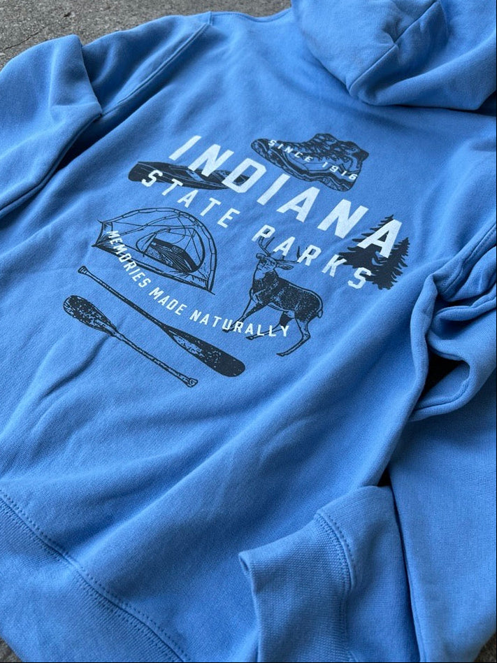 A light blue pullover hoodie laid flat on a gray asphalt surface, featuring a dark graphic print on the front that reads 'Indiana State Parks' with camping, hiking, and outdoor illustrations.