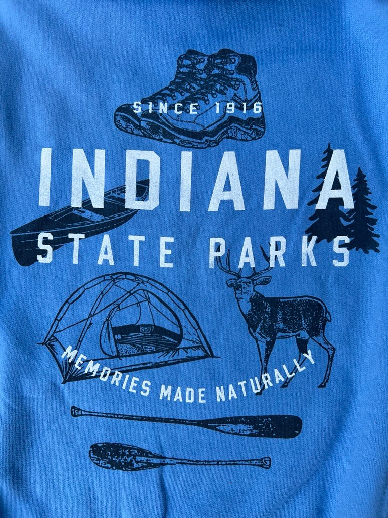 A close-up of a blue graphic t-shirt featuring 'INDIANA STATE PARKS' in white text, surrounded by outdoor illustrations including a hiking boot, canoe, tent, deer, canoe, and trees. The text 'SINCE 1916' and 'MEMORIES MADE NATURALLY' are also printed on the shirt.