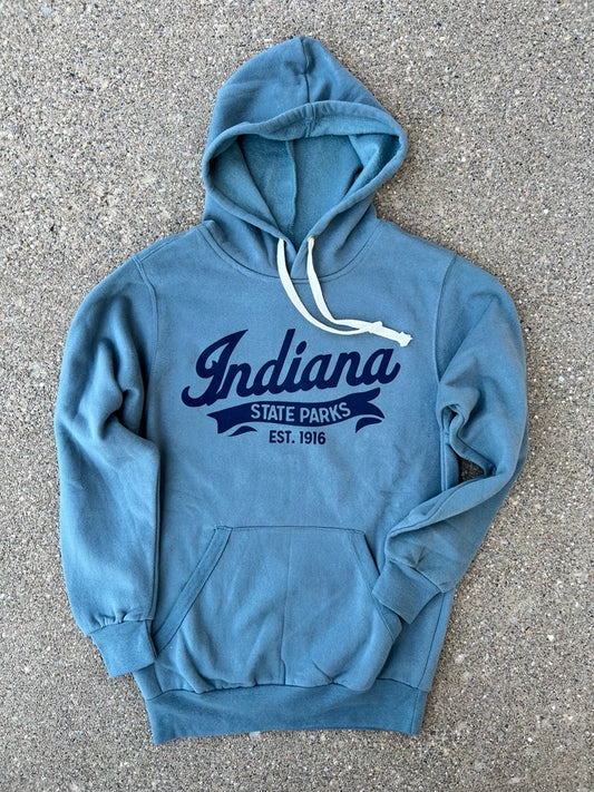 A slate blue pullover hoodie with white drawstrings and a navy blue graphic reading 'Indiana State Parks Est. 1916' on the front, laid flat on a gray asphalt surface.