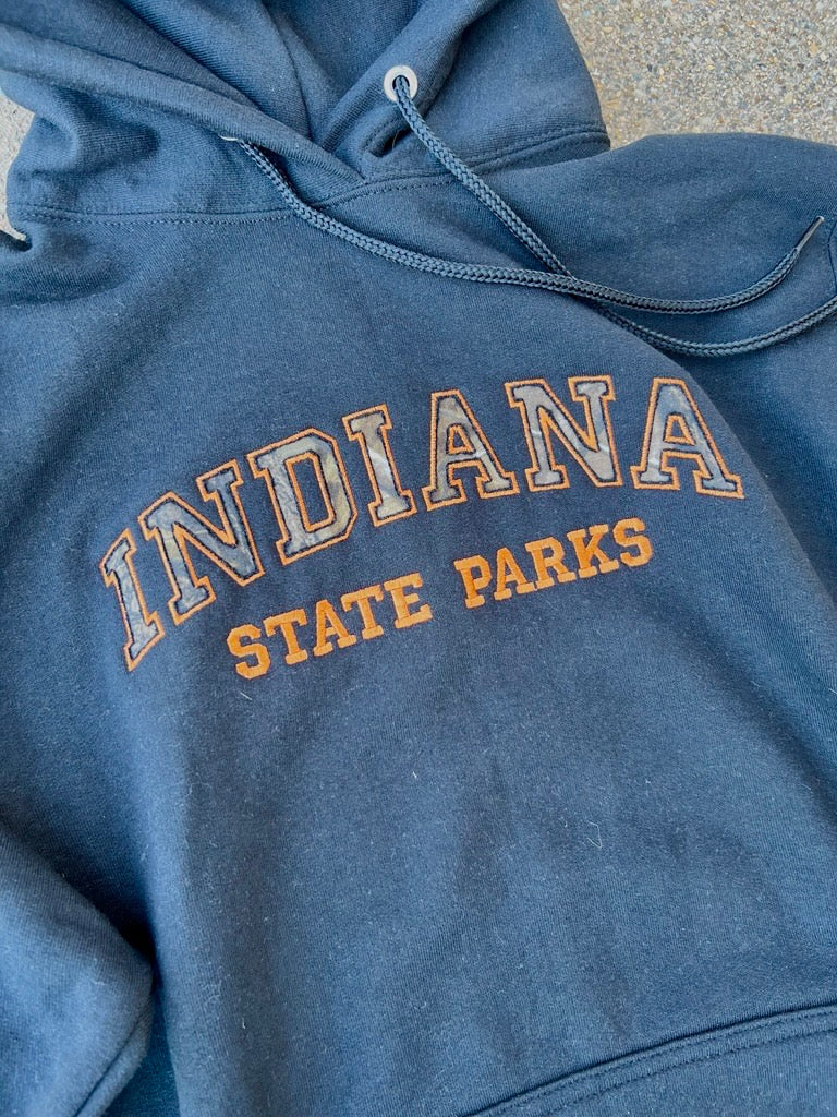 A close-up of a black hooded sweatshirt with 'INDIANA STATE PARKS' printed in orange and white varsity-style lettering on the chest.