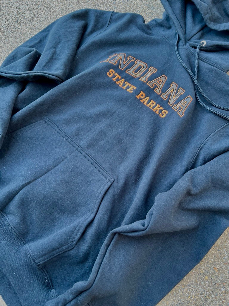 A black hooded sweatshirt laid flat on a gray asphalt surface, featuring 'INDIANA STATE PARKS' printed in tan and orange varsity lettering on the chest.