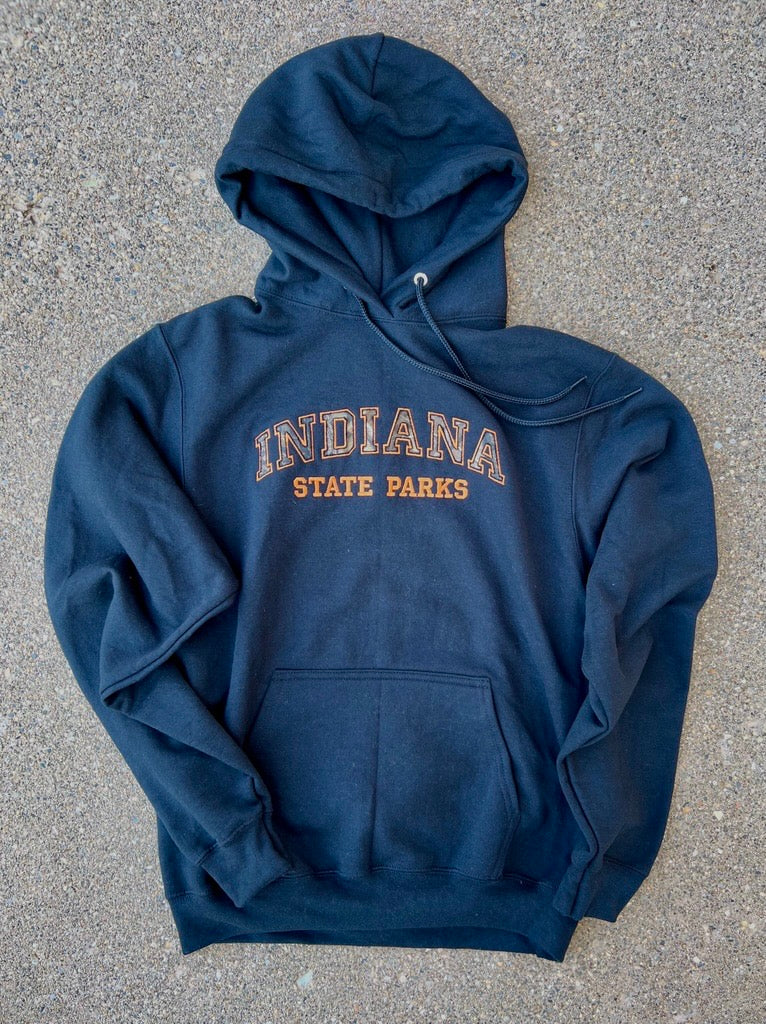 A black pullover hoodie with 'INDIANA STATE PARKS' printed in orange and camo on the chest, laid flat on a gray concrete surface.