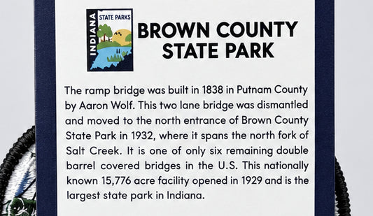 A close-up of a printed informational sign for Brown County State Park, Indiana, with the park logo at the top left and a block of text describing the ramp bridge and park history. Part of a dark blue border and a black rope are visible around the sign.