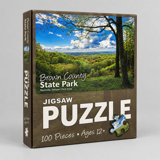 A boxed jigsaw puzzle package for Brown County State Park, showing a scenic landscape photo on the front with trees, blue sky, and green hills. The box is brown and reads 'JIGSAW PUZZLE,' '100 Pieces,' and 'Ages 12+'.