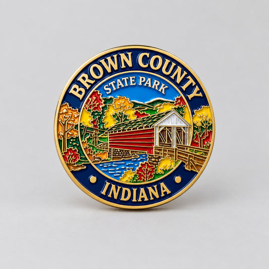 A round enamel pin featuring the Brown County State Park logo from Indiana, with a gold-tone border, blue outer ring, and a colorful central illustration of a covered bridge, autumn trees, and a river.