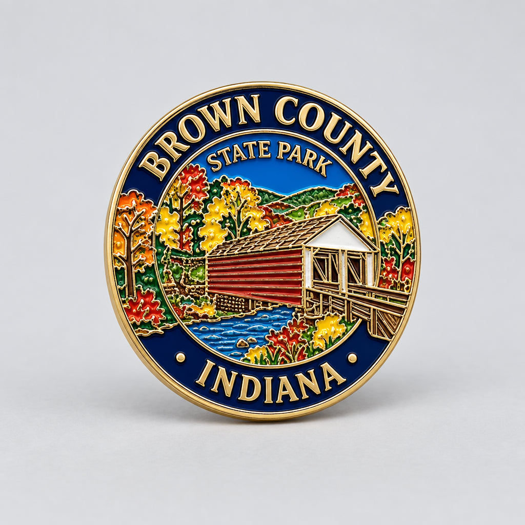 A round enamel pin featuring the Brown County State Park, Indiana design, with a gold-colored border, blue outer ring, and a colorful central scene depicting a covered bridge, autumn trees, and water.