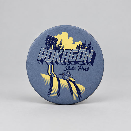 A round magnet showing a toboggan tower, evergreen trees, and two people riding a toboggan down icy tracks, with the text “Pokagon State Park.”