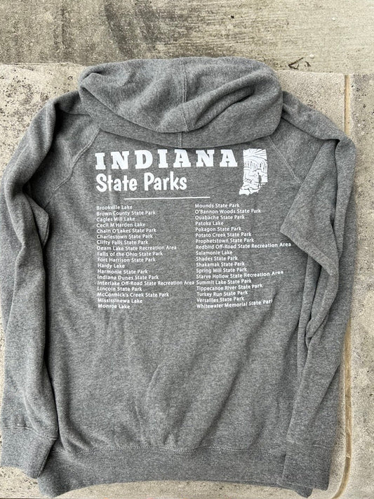 A heather gray hooded sweatshirt laid flat on a concrete surface, showing the back printed with 'INDIANA State Parks' and a list of parks. A person's pink shoes are partially visible at the bottom edge.