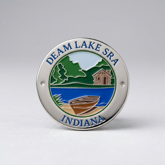 Round lapel pin with an enamel‑style scene showing a cabin, trees, hills, and a boat by the water, labeled “Deam Lake SRA Indiana.”