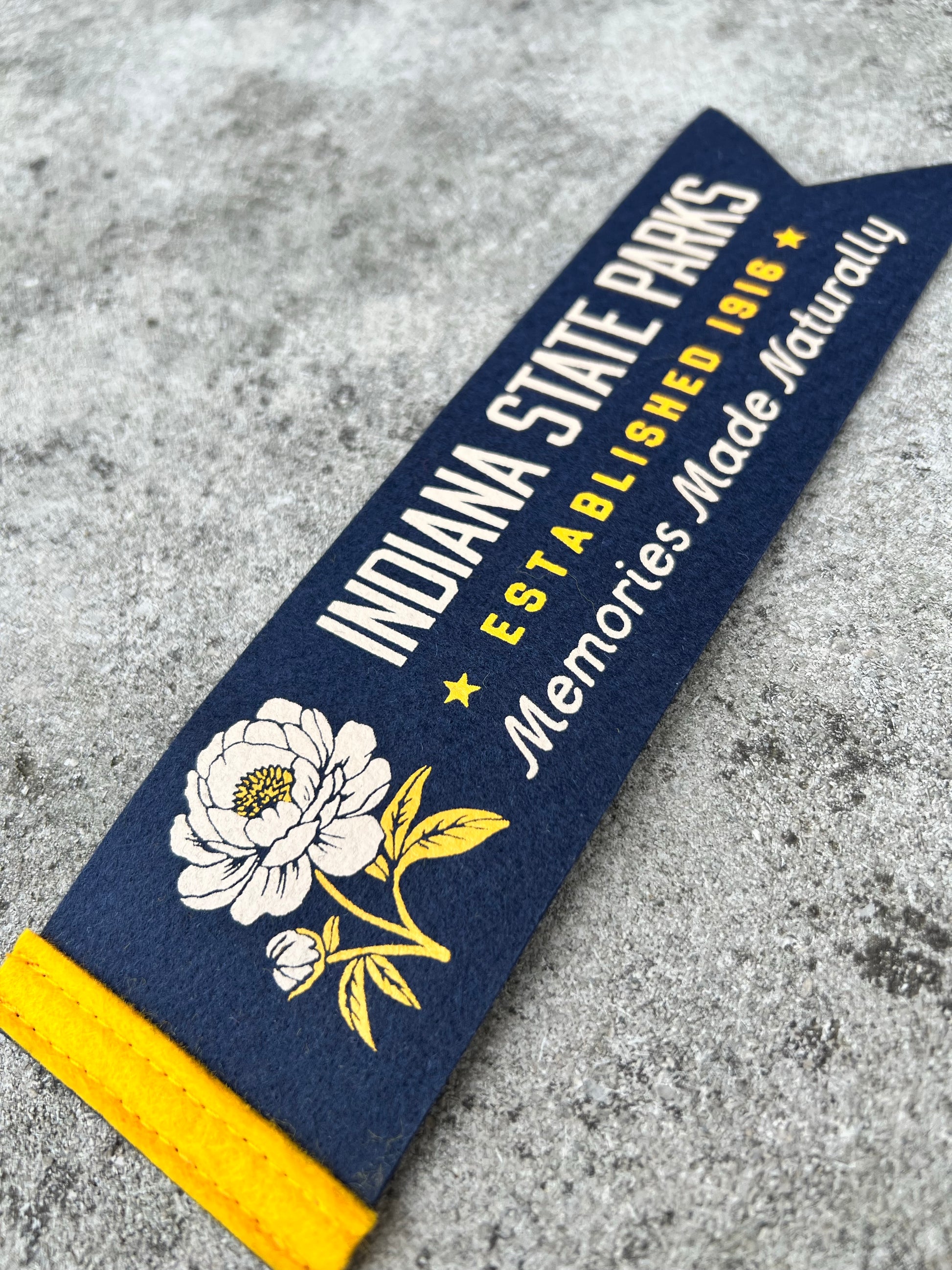 A navy blue felt pennant banner featuring 'INDIANA STATE PARKS ESTABLISHED 1916 Memories Made Naturally' in white and yellow text, decorated with a white peony illustration and a yellow end cap, shown on a gray concrete surface.