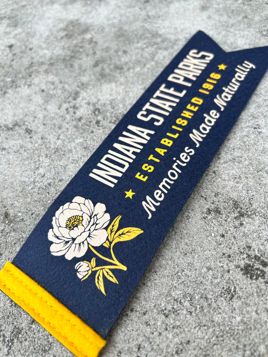 A navy blue felt pennant banner featuring 'INDIANA STATE PARKS ESTABLISHED 1916 Memories Made Naturally' in white and yellow text, decorated with a white peony illustration and a yellow end cap, shown on a gray concrete surface.