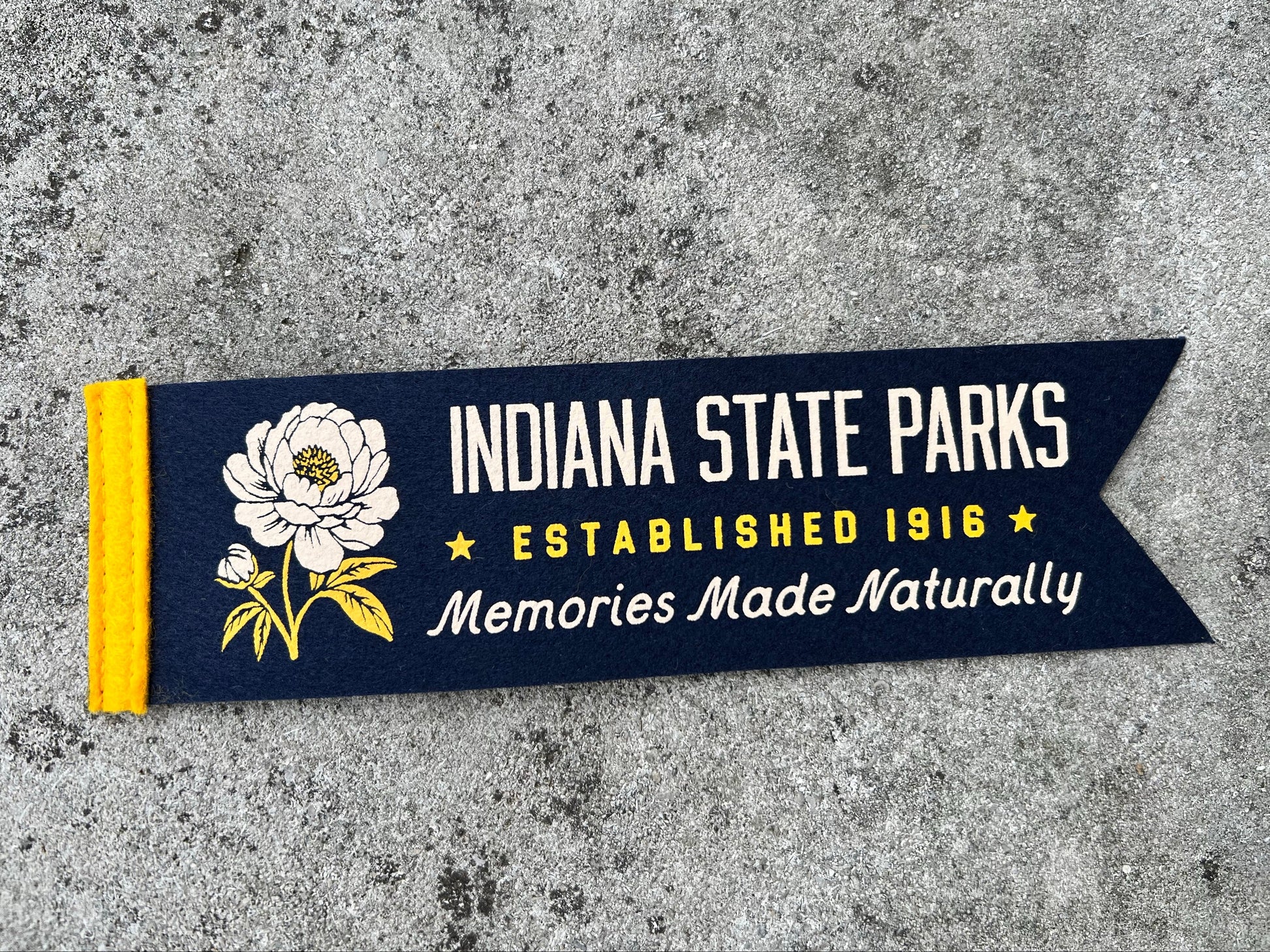 A navy blue fabric pennant banner featuring a white flower illustration and the text 'INDIANA STATE PARKS', 'ESTABLISHED 1916', and 'Memories Made Naturally', laid flat on a textured gray concrete surface.
