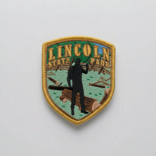 Embroidered shield‑shaped patch showing a silhouette figure with an axe among logs, labeled “Lincoln State Park.”