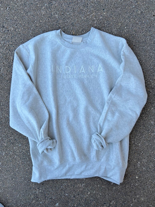 A light heather cream crewneck sweatshirt with 'INDIANA STATE PARKS' printed in a faded white tone across the chest, laid flat on a dark gray asphalt surface.