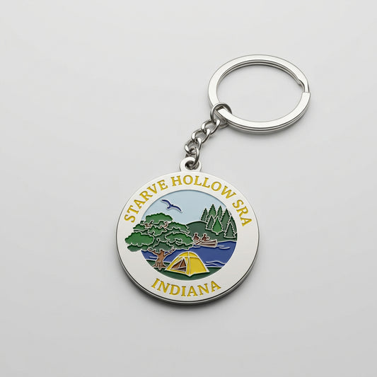 Round metal keychain with an enamel scene showing a tent, trees, water, and a flying bird, labeled “Starve Hollow SRA Indiana.”