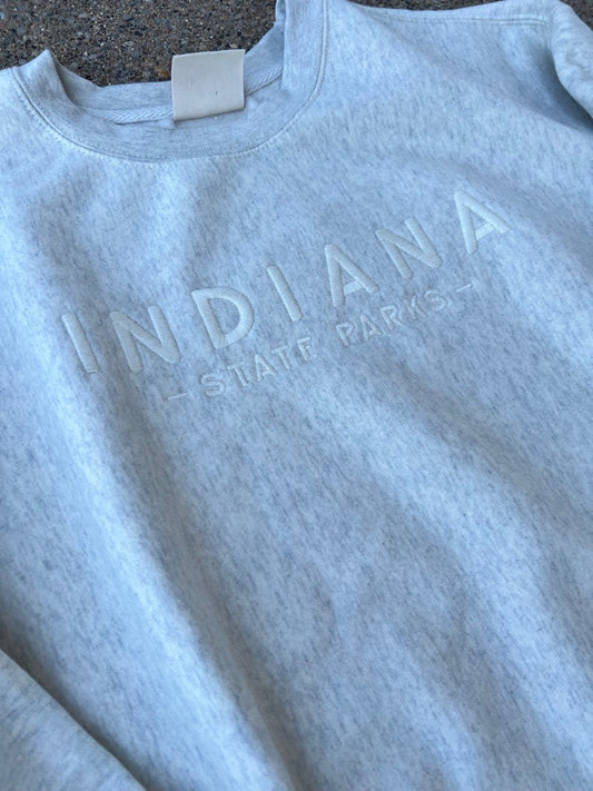 A close-up of a light heather cream sweatshirt featuring the words 'INDIANA STATE PARKS' in a tonal, embossed-style font across the chest.