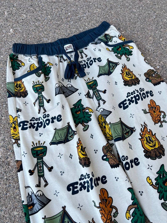 Close up of cream drawstring pj pants with a colorful cartoon camping-themed print featuring the phrase 'Let's Go Explore' repeated, laid flat on a gray asphalt surface.