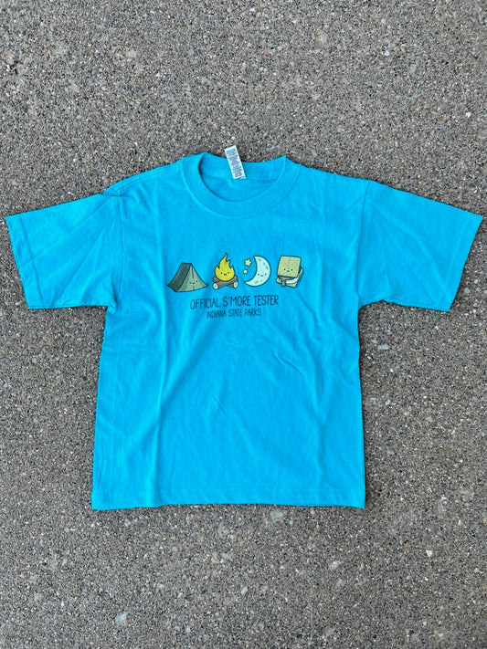 A bright turquoise blue short-sleeve youth t-shirt laid flat on a gray asphalt surface. The shirt features a small graphic on the chest showing a green tent, a yellow campfire, a white moon, and a brown s'more, with the text 'OFFICIAL S'MORE TESTER' and 'INDIANA STATE PARKS'.