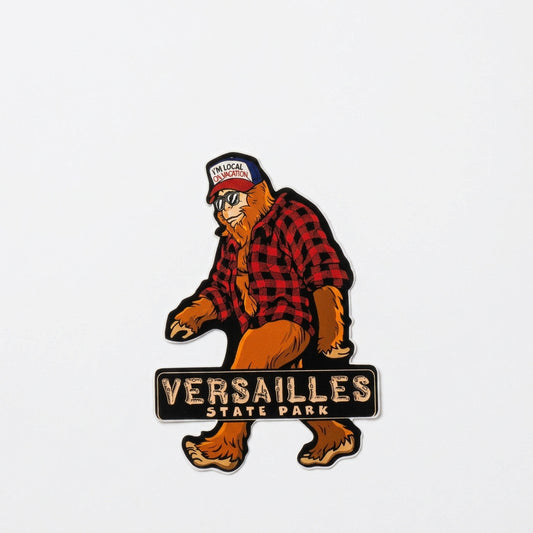 Die‑cut sticker of bigfoot walking while wearing a red plaid shirt and a hat that reads “I’M LOCAL ON VACATION,” with the text “Versailles State Park” below.
