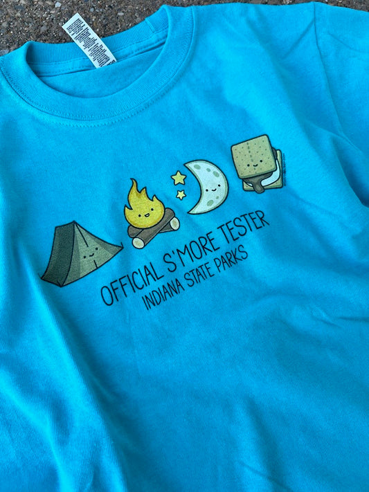 A bright turquoise blue youth t-shirt laid flat on a dark textured surface. The shirt features a cute cartoon graphic of camping and s'mores (tent, campfire, moon, s'more) with the text 'OFFICIAL S'MORE TESTER' and 'INDIANA STATE PARKS' printed below.