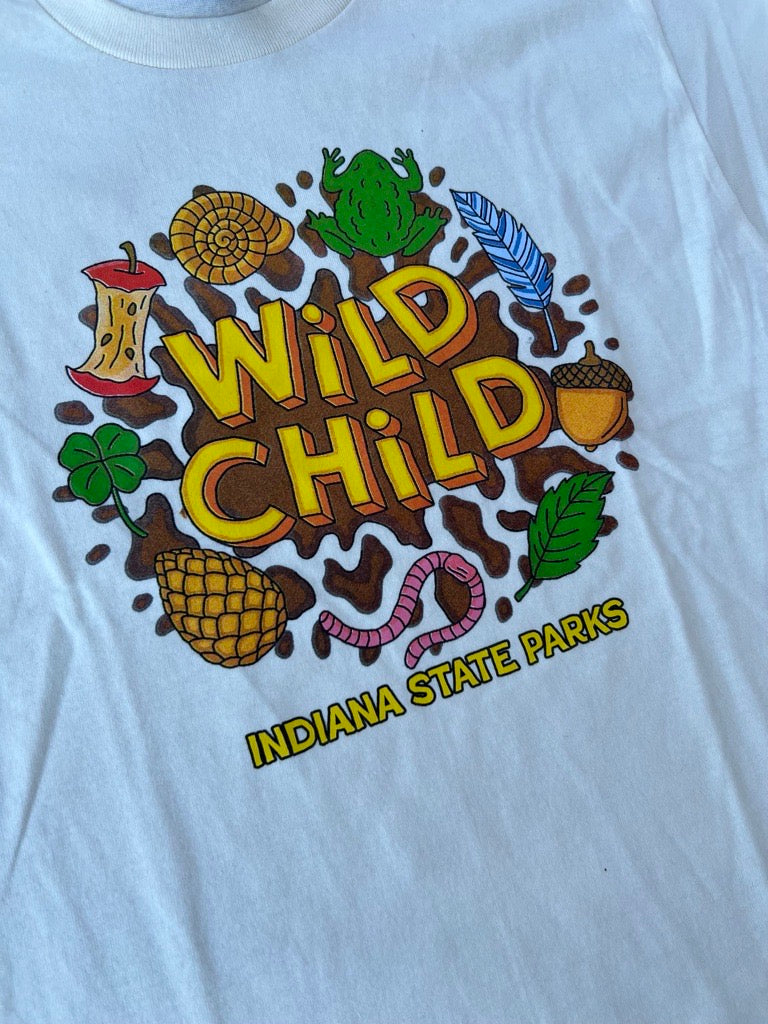 A close-up of a cream youth t-shirt featuring a colorful graphic print that reads 'Wild Child' with 'Indiana State Parks' below it. The graphic includes illustrations of a green frog, pinecone, acorn, worm, and leaves.