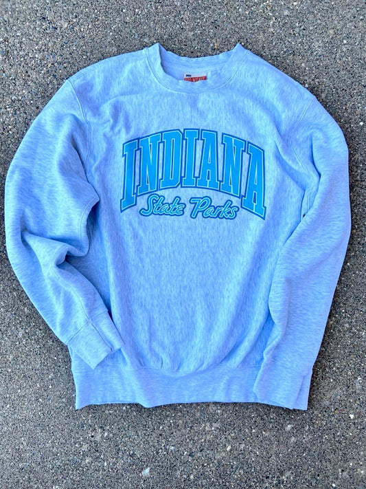 A light gray heather crewneck sweatshirt with 'INDIANA State Parks' printed in blue on the front, laid flat on gray asphalt.