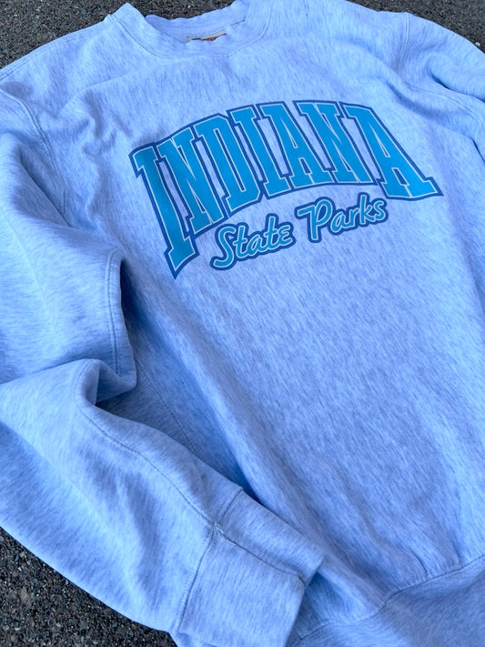 A light gray crewneck sweatshirt featuring 'INDIANA State Parks' printed in blue arched lettering across the chest, laid flat on a dark asphalt surface.