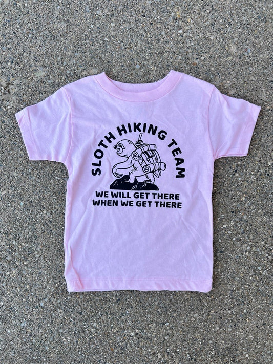 A light pink short-sleeve toddler t-shirt with a black graphic of a sloth carrying a hiking pack, surrounded by the text 'SLOTH HIKING TEAM' and 'WE WILL GET THERE WHEN WE GET THERE'. The shirt is laid flat on a gray concrete surface.