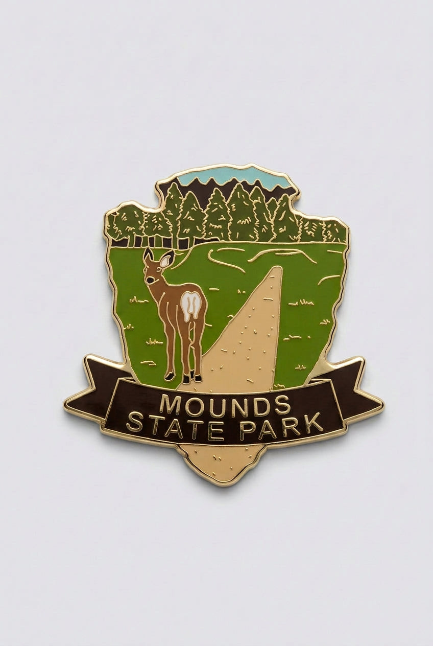 Enamel‑style magnet with a deer standing on a forest trail, framed by trees and a sky background, labeled “Mounds State Park.”