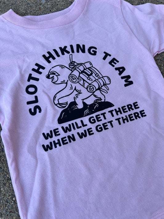 A light pink short-sleeve toddler t-shirt laid flat on a gray concrete surface, featuring a black graphic of a sloth hiking with the text 'SLOTH HIKING TEAM' and 'WE WILL GET THERE WHEN WE GET THERE'.