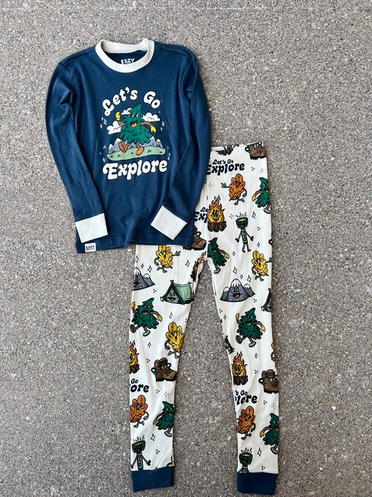 A two-piece children's pajama set laid flat on gray pavement. The long-sleeve top is navy blue with a white collar and cuffs, featuring a green tree graphic and the words 'Let's Go Explore.  The pants are cream colored with camping graphics.